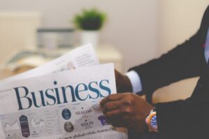 A person in a suit reads a newspaper with the headline "Business" in a modern office setting.