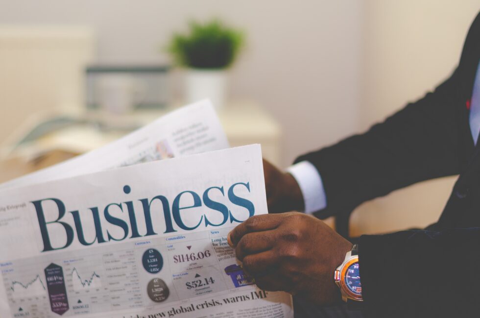 A person in a suit reads a newspaper with the headline "Business" in a modern office setting.