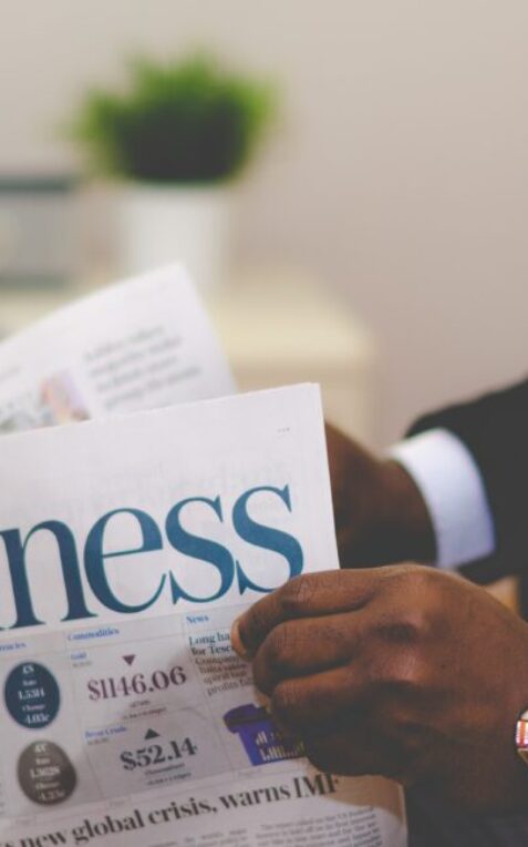 A person in a suit reads a newspaper with the headline "Business" in a modern office setting.