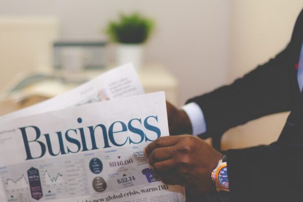 A person in a suit reads a newspaper with the headline "Business" in a modern office setting.